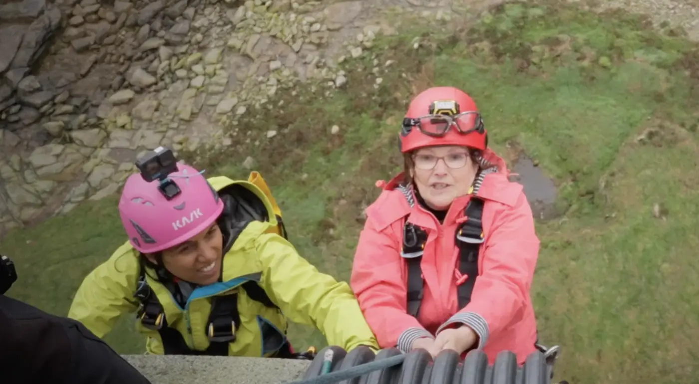 Deux challengeuses souriantes, l'une en casque rose, l'autre en casque rouge, se hissent sur un rocher herbeux.