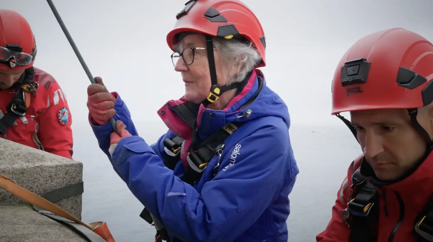 Une challengeuse concentrée, casque rouge et veste bleue, sur le phare de l'Île Vierge, à côté d'un secouriste en tenue rouge.