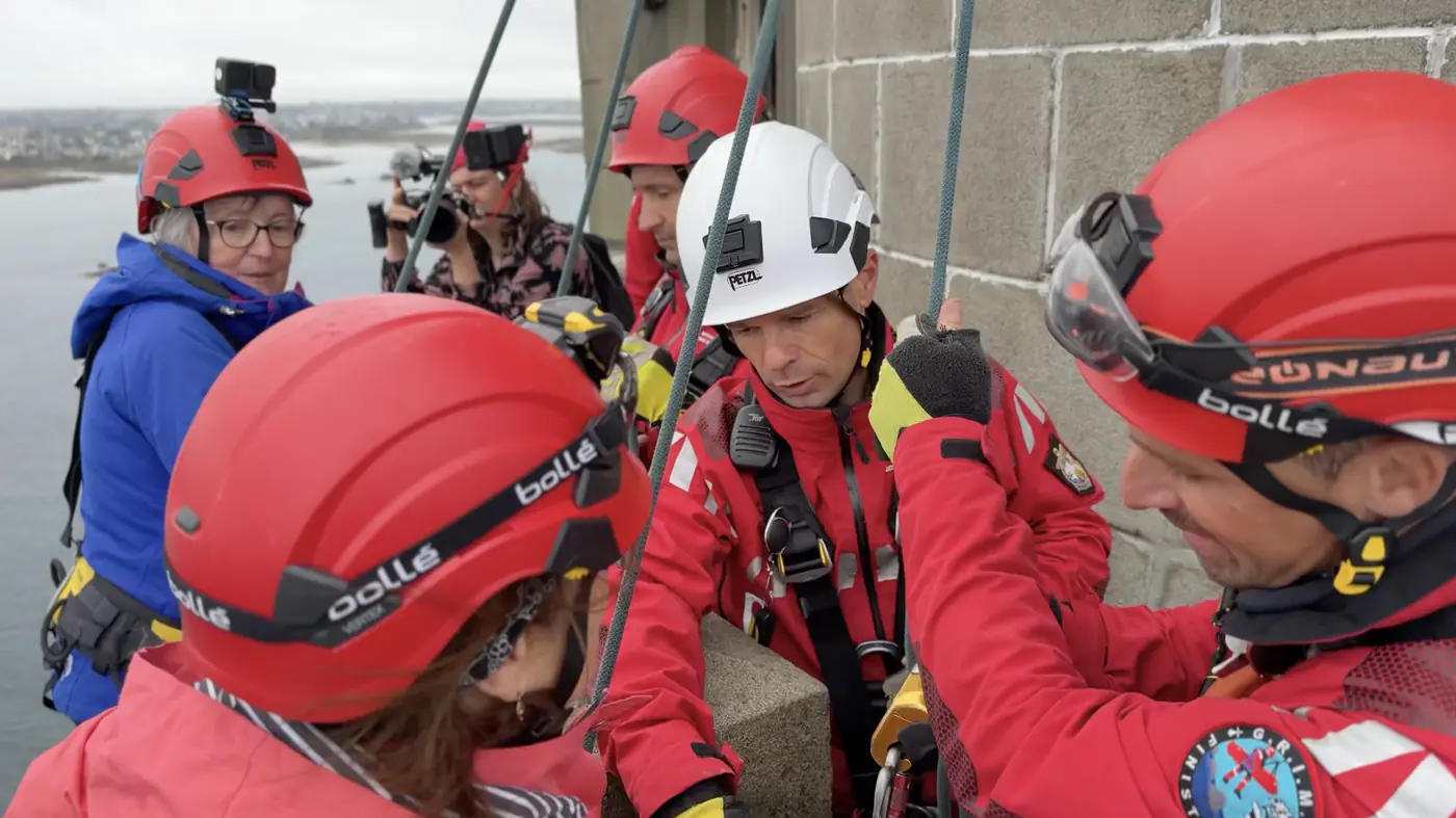 Une équipe de secouristes en tenue rouge et casques ajuste le matériel de rappel au sommet du phare.